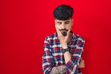 Young hispanic man with beard standing over red background thinking looking tired and bored with depression problems with crossed arms.