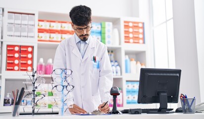 Young hispanic man pharmacist writing on paper standing at pharmacy