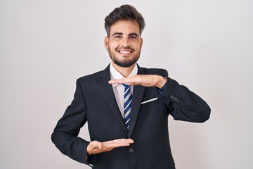 Young hispanic man with tattoos wearing business suit and tie gesturing with hands showing big and large size sign, measure symbol. smiling looking at the camera. measuring concept.
