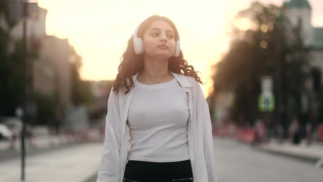 Charming young indian woman walking down the street with headphones listening to music and looking around at evening outdoors Happy relaxed lady walking on the city centre enjoying beautiful sunset