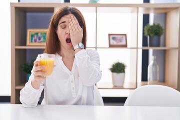 Brunette woman drinking glass of orange juice yawning tired covering half face, eye and mouth with...