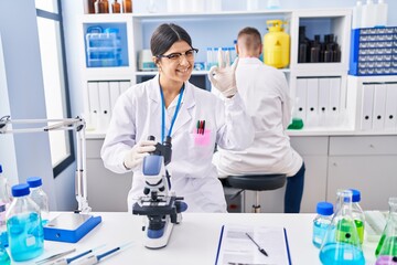 Fototapeta premium Young woman working at scientist laboratory doing ok sign with fingers, smiling friendly gesturing excellent symbol