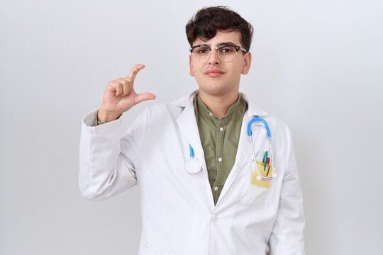 Young non binary man wearing doctor uniform and stethoscope smiling and confident gesturing with hand doing small size sign with fingers looking and the camera. measure concept.