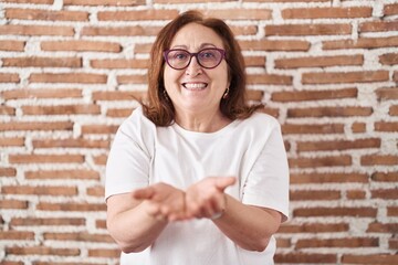 Senior woman with glasses standing over bricks wall smiling with hands palms together receiving or...