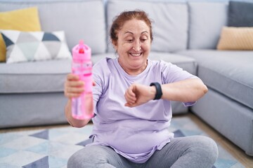 Fototapeta premium Senior woman sitting on floor drinking water looking watch at home