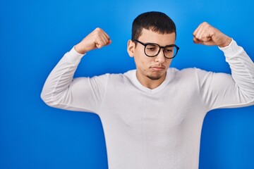 Young arab man wearing casual white shirt and glasses showing arms muscles smiling proud. fitness concept.