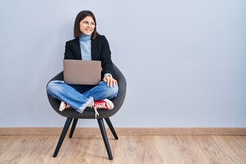 Young hispanic woman sitting on chair using computer laptop looking away to side with smile on...