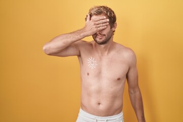 Fototapeta premium Caucasian man standing shirtless wearing sun screen covering eyes with hand, looking serious and sad. sightless, hiding and rejection concept