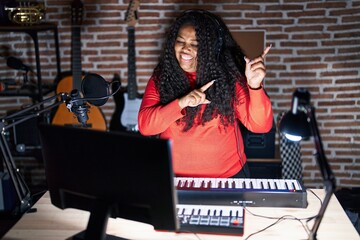 Plus size hispanic woman playing piano at music studio smiling and looking at the camera pointing with two hands and fingers to the side.
