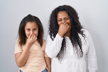 Mother and young daughter standing over white background smelling something stinky and disgusting,...