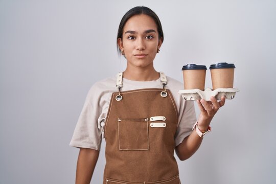 Young hispanic woman wearing professional waitress apron holding coffee relaxed with serious expression on face. simple and natural looking at the camera.