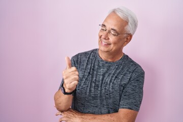 Middle age man with grey hair standing over pink background looking proud, smiling doing thumbs up gesture to the side