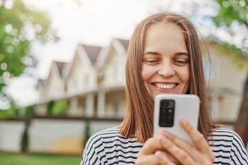Smiling brown haired woman browsing internet on smart phone standing outdoor in street with houses on background checking social network while walking outside.