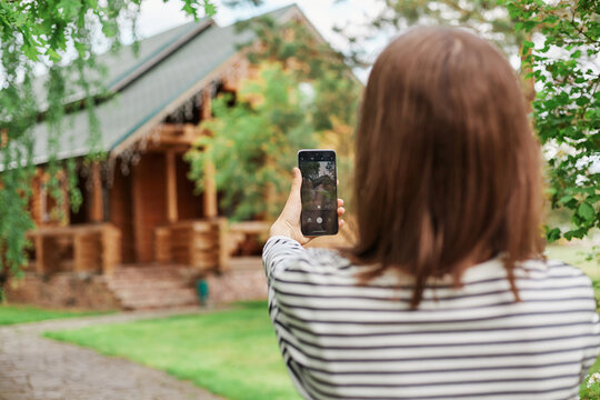 Back view of dark haired woman taking photo on her mobile phone wearing striped casual shirt holding smartphone in hands broadcasting livestream.