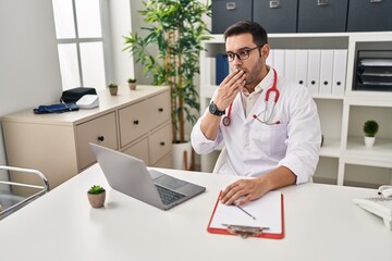 Young hispanic doctor man with beard doing online appointment at the clinic covering mouth with...
