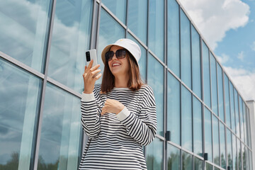 Joyful smiling woman wearing striped shirt using phone posing near building with big windows making selfie on her cell phone communicating with subscribers.