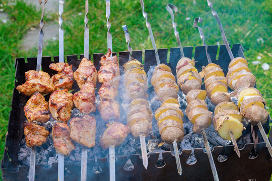 An array of kebabs skewers and potatoes topped with bacon are cooked on portable metal BBQ braziers