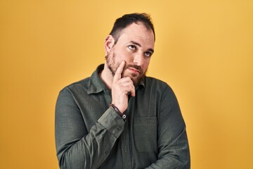 Plus size hispanic man with beard standing over yellow background with hand on chin thinking about question, pensive expression. smiling with thoughtful face. doubt concept.