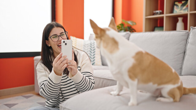 Young hispanic woman with chihuahua dog sitting on the floor taking photo with smartphone at home