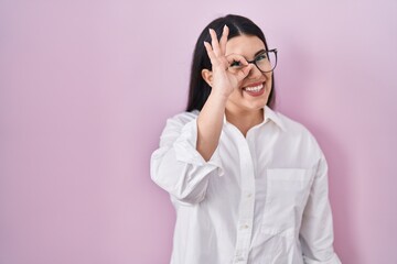 Young brunette woman standing over pink background doing ok gesture with hand smiling, eye looking through fingers with happy face.
