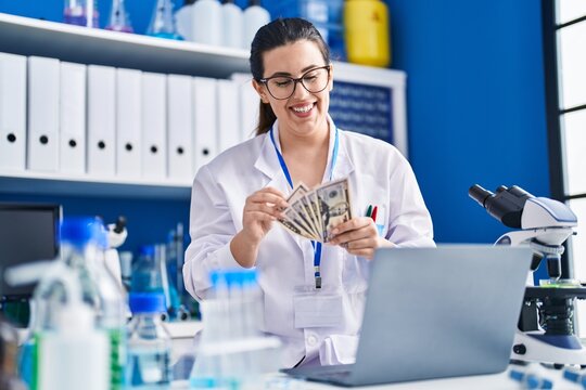 Young hispanic woman scientist using laptop counting dollars at laboratory