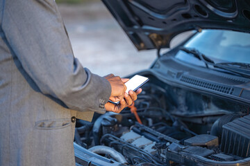 Detail of the hands of a Latino man with his mobile phone with his broken down car in the background.
