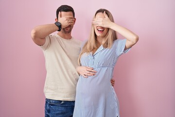 Young couple expecting a baby standing over pink background smiling and laughing with hand on face covering eyes for surprise. blind concept.