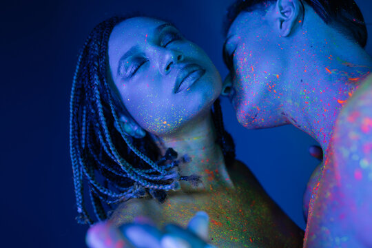 sexy interracial couple in colorful neon body paint, african american woman with dreadlocks and closed eyes near young bare-chested man on blue background with cyan lighting