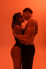 Naklejka premium side view of interracial passionate couple, shirtless man in black pants with muscular torso and sexy african american woman in lace bodysuit embracing on orange background with red lighting effect