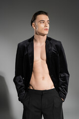 Fototapeta premium young, sexy and fashionable man wearing black silk blazer on shirtless muscular torso and looking away while posing with hands in pockets of pants on grey background