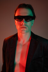 portrait of young and fashionable man in dark sunglasses and stylish blazer looking at camera while standing and posing on grey background with red lighting