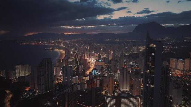 Big coastal city next to the calm blue ocean at night. Pink sunset and dark blue clouds in the background. Bright city lights and busy night life. Aerial view. 