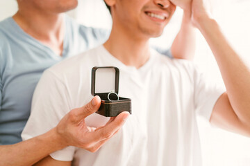 Young happy gay couple in love, proposing a marriage surprise with a wedding ring in the living room at home. Gay men proposal engagement concept. Surprised partner is very happy. Relationship goal.