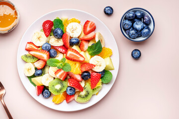 Summer healthy fruit and berry salad with strawberries, blueberries, banana, kiwi, orange and mint leaves, pink background, top view