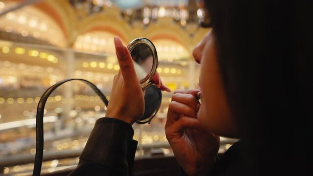 Beautiful young woman applying lipstick looking at a small mirror, at the Galeries Lafayette, in Paris, France