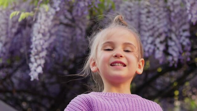 Cute girl in a lilac sweater against the background of blooming wisteria laughs merrily, children's portrait of a preschooler, Children's Day