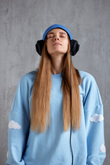A young pretty long-haired girl in a blue sweater, a funny hat and black headphones listens to music with her eyes closed. Meditation and immersion in music. Half-length studio shot, gray background.