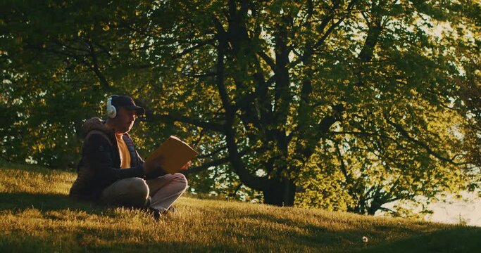 Young Man is Reading a Book in a Clearing in the Park With Headphones on His Head. Student Studying from a Book Sitting Grass in a Public Park Listening to Music with Headphones and Reading a Book. 