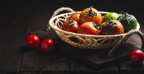 Still life of fresh colorful ripe fall or summer heirloom variety tomatoes in a basket on dark wooden background.