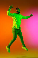 Fototapeta na wymiar Full-length portrait of handsome bearded man in casual clothes jumping against gradient studio background in neon light. Concept of human emotions, facial expression, lifestyle, fashion