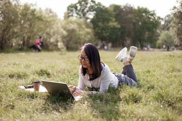 Naklejka na ściany i meble A asian woman is sitting on the grass in the park, working on a laptop. A woman with a laptop while sitting under a tree in a park with bright sunlight from behind.