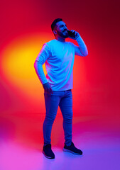 Portrait of handsome, smiling, bearded man in casual clothes talking on mobile phone against gradient studio background in neon light. Concept of human emotions, facial expression, lifestyle