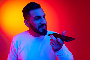Portrait of handsome bearded man in casual clothes smiling and recording voice on mobile phone against gradient studio background in neon light. Concept of human emotions, facial expression, lifestyle