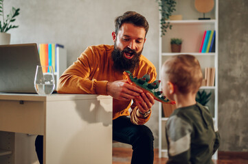 A cheerful bearded hipster is looking at the dinosaur toy with interest while sitting at his cozy...