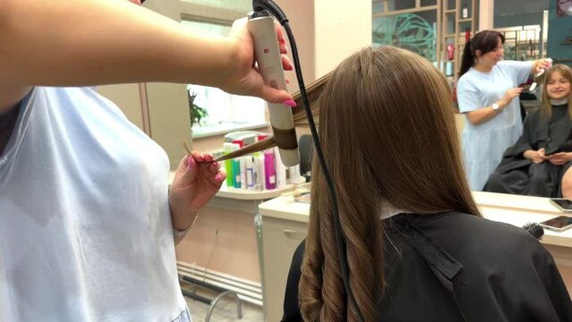 a female hairdresser makes a hairstyle for a girl in a beauty salon or a barber shop, a child's hairstyle for a child on a holiday, curls with a curler