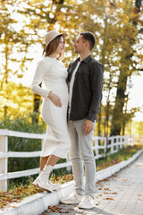 Fototapeta premium Pregnancy and Parenthoods concept - smiling man is walking with pregnant wife and looking to each other in autumn park. Happy styling young family is relaxing outdoors on weekend on sunny day.