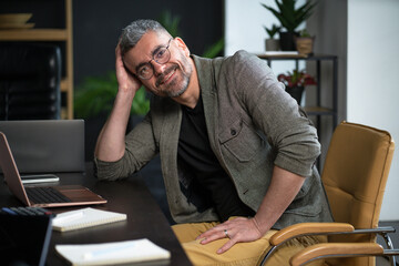 Happy worker sits in loft office, smiling with contentment work. Desk with computer, stationary,...