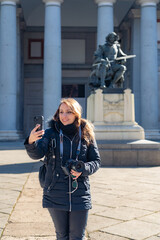 Smiling young Latin woman in warm clothes with professional photo camera taking selfie on smartphone while standing near Prado Museum in Madrid, Spain
