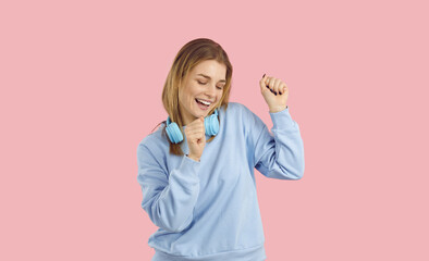Carefree young woman enjoying favorite song, dancing and feeling free and happy. Beautiful girl wearing trendy blue sweatshirt listening to music and dancing isolated on solid pastel pink background