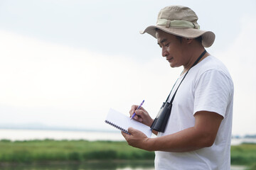 Asian man ecologist is surveying nature at the lake, write on paper notebook. Concept, nature...